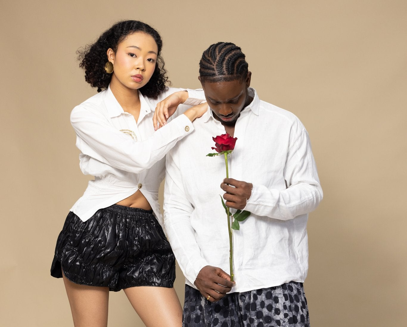 A man and a woman posing together against a solid beige studio background. The woman wears a white button-down shirt and black patterned shorts, while the man wears a white button-down shirt and a dark patterned long-sleeve top, holding a single red rose.