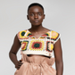 A model wearing a sleeveless crochet crop top with a multicolor granny square pattern and matching high-waisted peach shorts, set against a plain light gray studio background.