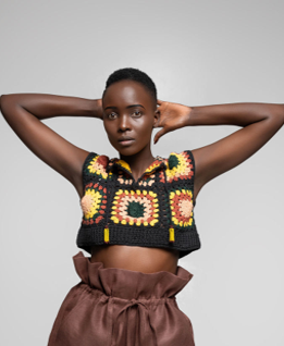 A model posing in a black crochet crop top with colorful sunflower patterns and brown paperbag shorts against a light gray studio background.